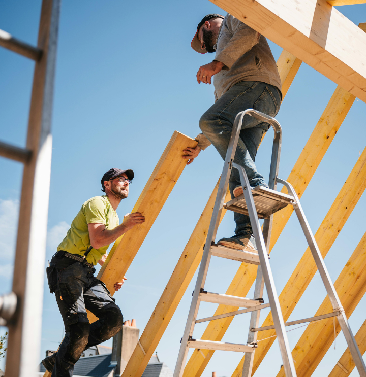 Two Roofers building a roof, one on a ladder and the other passing a piece of wood across.
